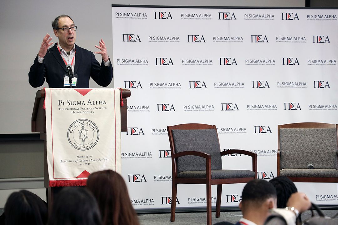 Matt Dallek speaking at a podium, next to a Pi Sigma Alpha Student Conference step-and-repeat