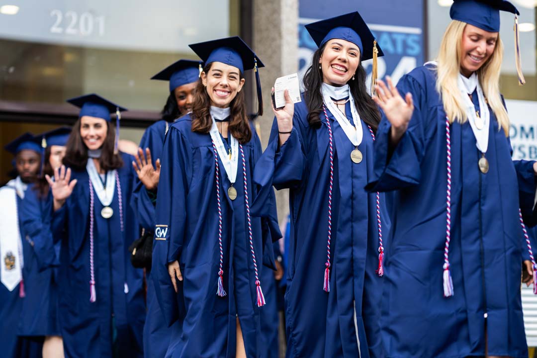 Students in cap and gowns wave to camera as they proceed to graduation.