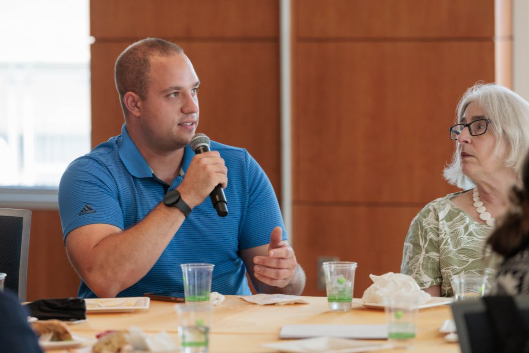 A man sitting down at a large table speaks into a microphone