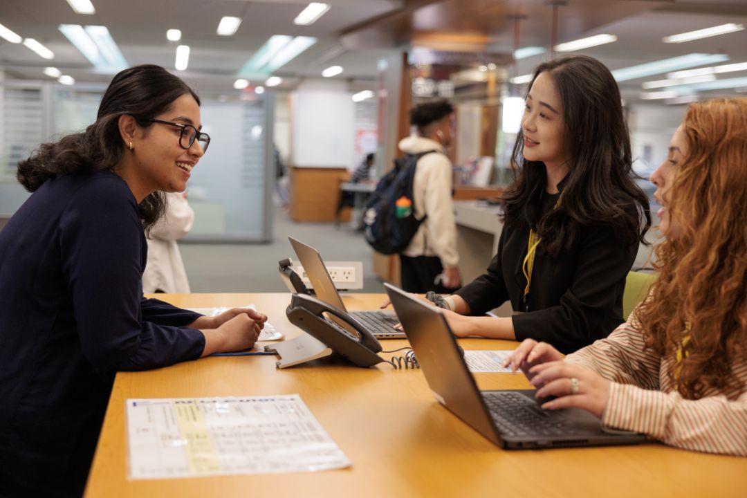 Three students sit at a table in the library with their laptops