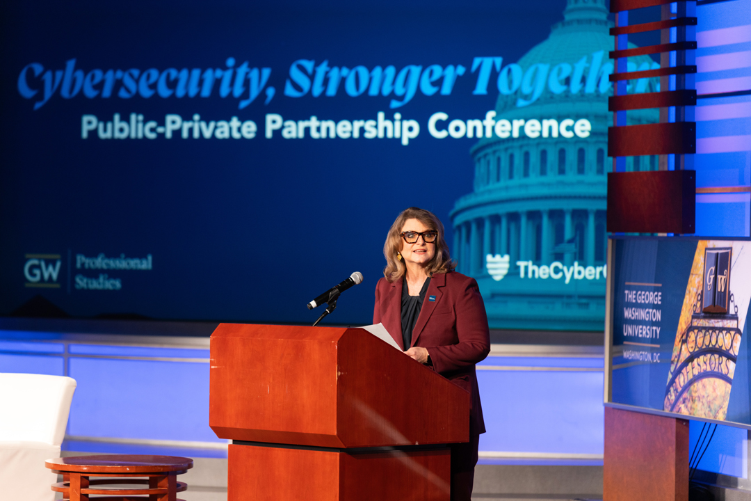 Liesl Riddle standing at a podium onstage in front of a screen that reads "Cybersecurity, Stronger Together. Public-Private Partnership Conference