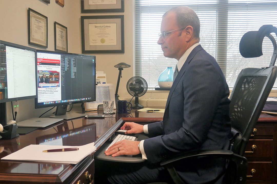 Dr. Todd Belt in a dark suit, white shirt and glasses in his office looking at a computer monitor watching Michael Cohen testify