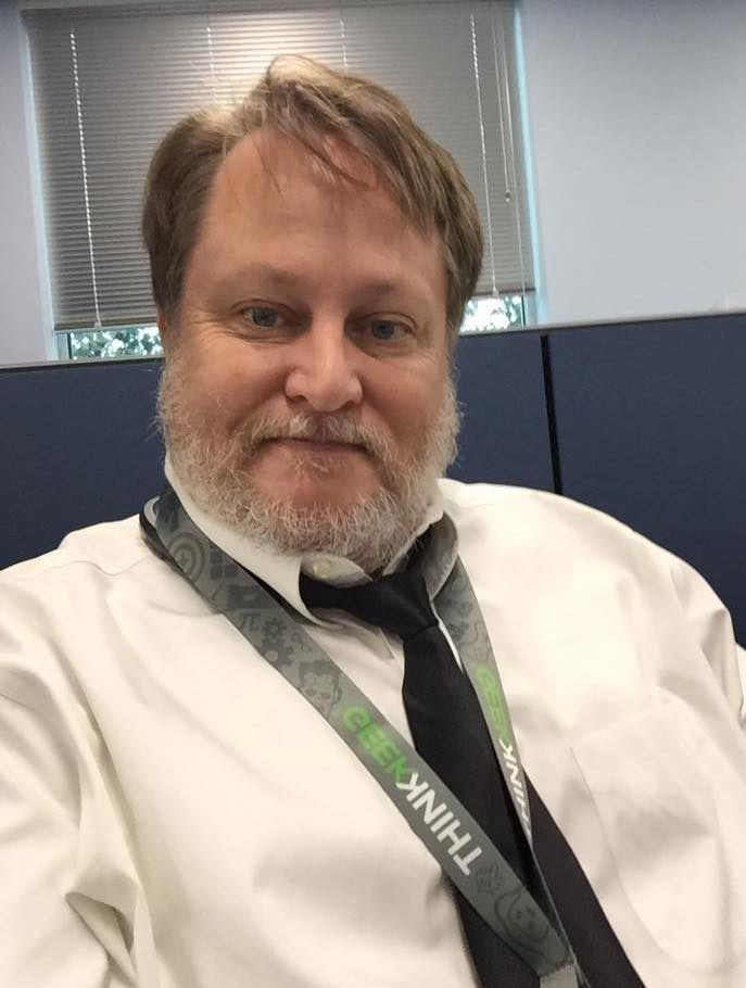 Todd Blanchard with grey beard and hair combed long with button down white shirt, tied and lanyard