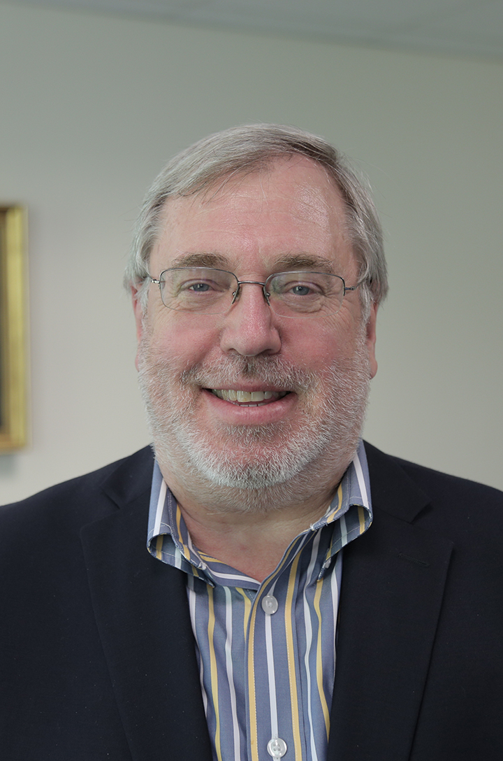 Man with glasses short hair and beard in navy blazer and striped collared shirt smiling