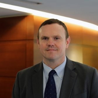 Man with brown hair, suit and tie in front of shiny wooden golden paneled wall with light reflecting