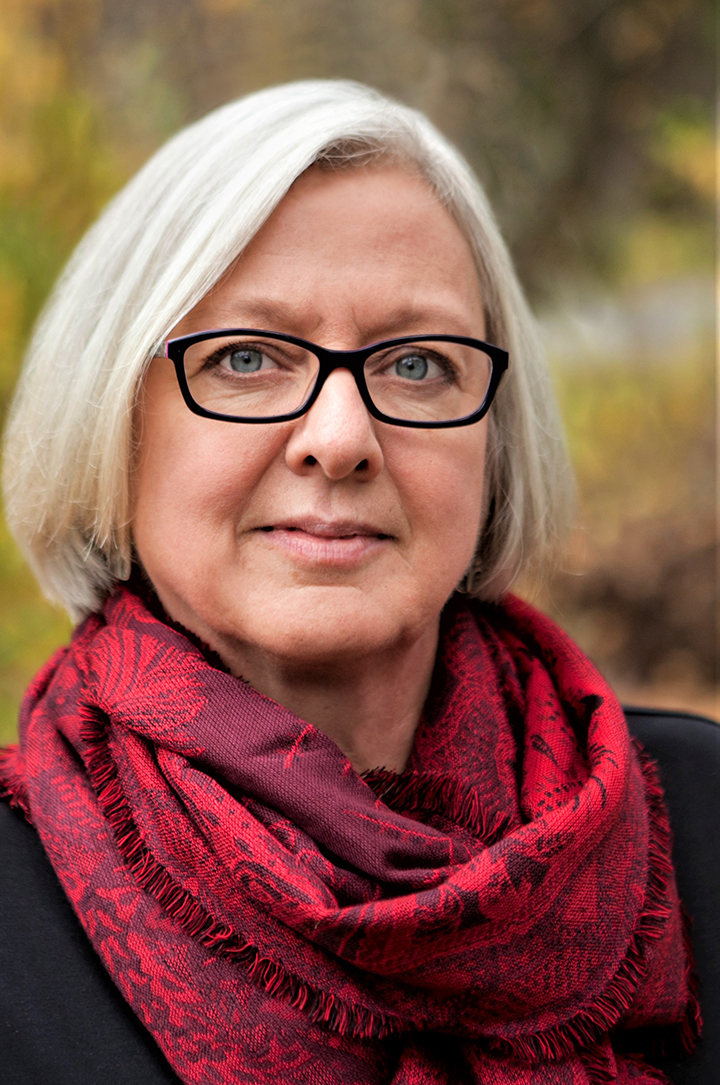 headshot of woman with grey bob, glasses, red patterned scarf and multi colored background that looks like autumn leaves
