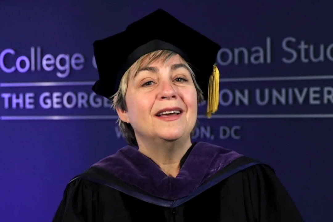 Toni Marsh wearing commencement gown and hat with tassel and part of College of Professional studies logo on a blue wall behind 