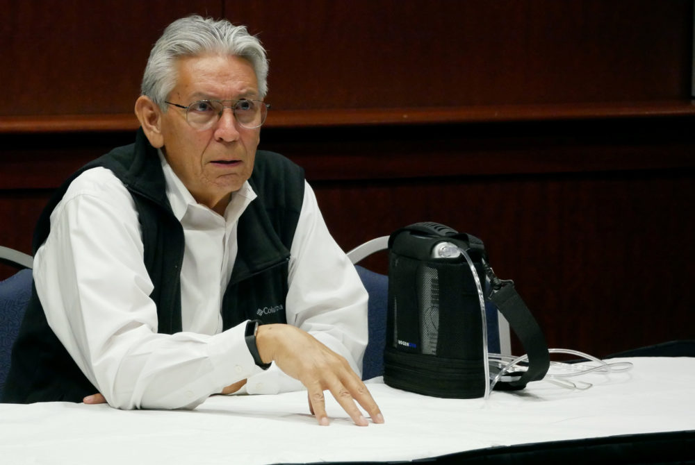 Kevin Gover speaks at GW, sitting at a table, white shirt, black vest, glasses