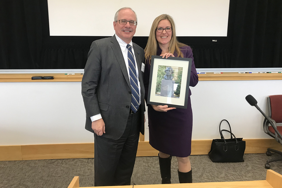 U.S. Rep. Wexton, professional attire, long blond hair, stands to the right of GW Pres. LeBlanc in suit. She is holding a photo.