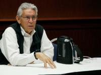 Kevin Gover speaks at GW, sitting at a table, white shirt, black vest, glasses