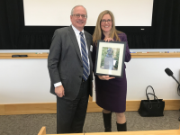 U.S. Rep. Wexton, professional attire, long blond hair, stands to the right of GW Pres. LeBlanc in suit. She is holding a photo.