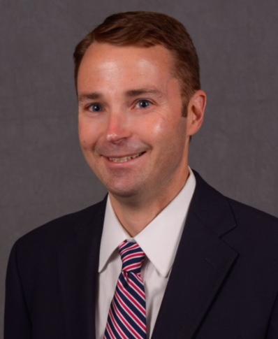 Gannon smiling youthful man with dark suit, white collared shirt, striped red/blue tie, short brown hair