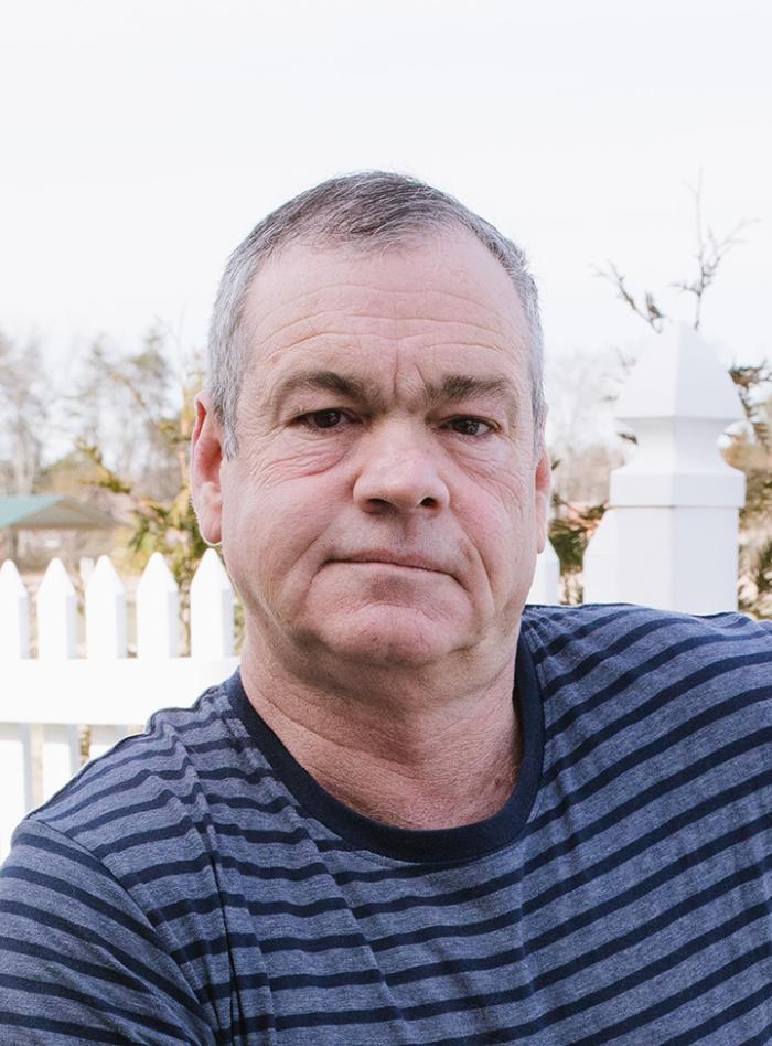 Man with short grey hair and striped shirt with shades of blue and white fence and foliage behind him