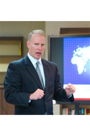 John Kavanagh teaching in a suit at GW with a monitor with a map on it