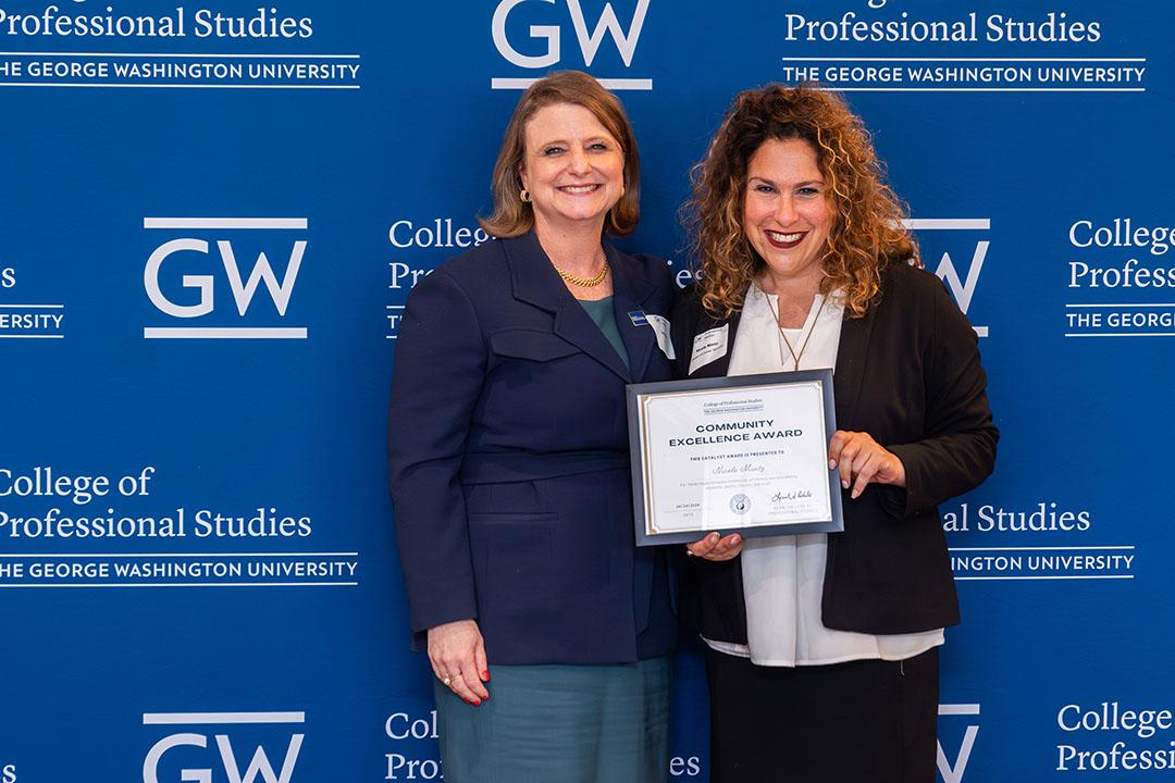 Two women with award frame in front of a GW blue background with logos